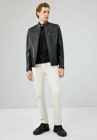 Zwarte leren jas met een staande kraag, twee met rits gesloten kleurzakken aan de voorkant, gecombineerd met een zwart shirt en witte broek. Zwarte sneakers maken de look compleet.