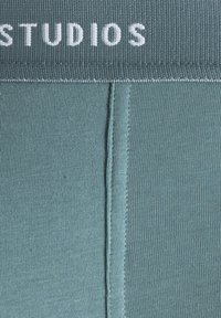 Tight-fitting teal leggings with a ribbed texture and a solid waistband featuring the word "STUDIOS" in white uppercase letters.