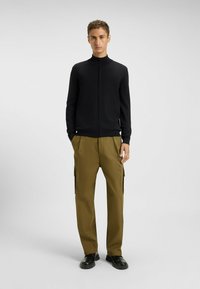 Black zip-up sweater with a mock neck, paired with olive cargo pants featuring side pockets. Model wears black shoes.