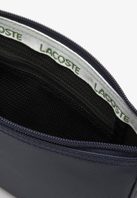 Pochette zippée marine avec un extérieur texturé, un intérieur doublé noir et un bord avec le logo blanc de Lacoste à l'ouverture. Dotée d'une fermeture éclair lisse.