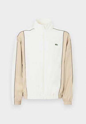 Giacca bianca con zip, maniche in tessuto beige, rifiniture nere e un piccolo logo verde sul petto. Presenta polsini e orlo elastici.