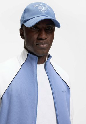 Mann trägt hellblaue und weiße Reißverschlussjacke und passende blaue Kappe mit Tennisclub-Logo auf weißem Hintergrund.