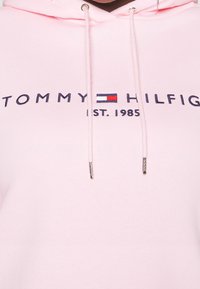 Vaaleanpunainen huppari, jossa on kiristettävä huppu ja "TOMMY HILFIGER" -logo sekä perustamisvuosi tummansinisinä, jossa raidalliset logo-yksityiskohdat.