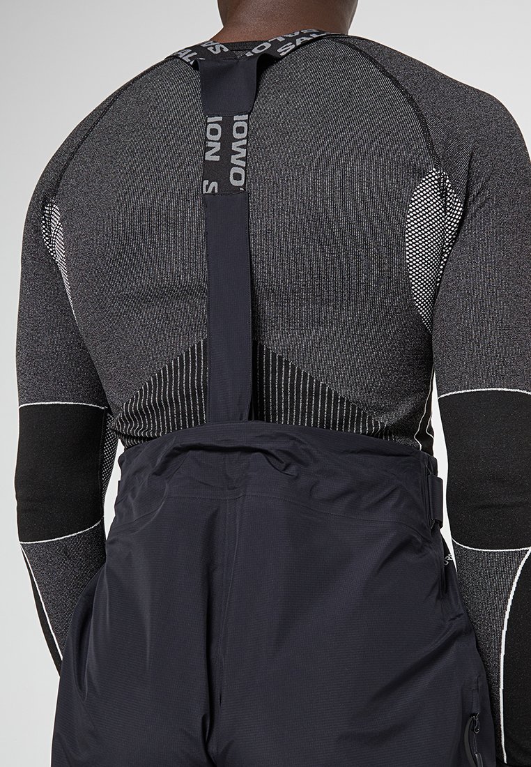 Fijne, gestructureerde zwarte basislaag met geometrische patronen en witte accenten, gecombineerd met donkere, verstelbare overall met ritszakken.