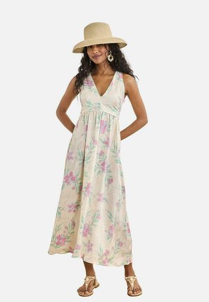 Mujer con vestido maxi beige sin mangas con estampado floral rosa, sombrero de paja de ala ancha, grandes pendientes y sandalias beige, con las manos detrás de la espalda.