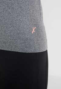 Grå kortärmad skjorta med ett strukturerat tyg och en rosa 'X'-logotyp nederst till vänster. Kombinerad med svarta leggings med en slät yta.