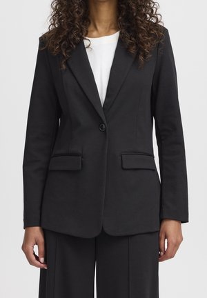 Frau trägt schwarzen maßgeschneiderten Blazer über weißem Top, mit passenden schwarzen Hosen, vor neutralem Hintergrund stehend.