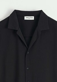 Chemise noire à col avec des boutons sur le devant et une étiquette blanche de taille S portant l'inscription "FRILIVIN" à l'intérieur du col.