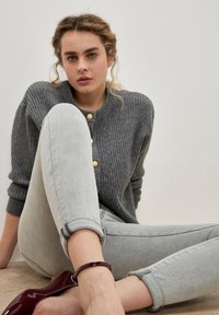 Joven mujer sentada en el suelo con las piernas cruzadas, vistiendo un cardigan de canalé gris con botones dorados, jeans claros con puños grises y zapatos de tobillo burdeos con tira.