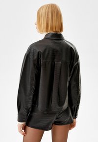 Czarna koszula z eko skóry z fakturą wzoru krokodyla, mankietami na guziki i nieco luźniejszym krojem z zaokrąglonym dołem.