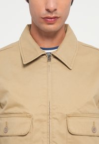 Tan jacket with a zippered front, pointed collar, and two chest pockets. Fabric appears smooth with a slight sheen.