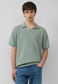 Polo à manches courtes, de couleur vert clair, en tissu texturé avec un décolleté en V et un col contrasté. Porté avec un jean bleu.