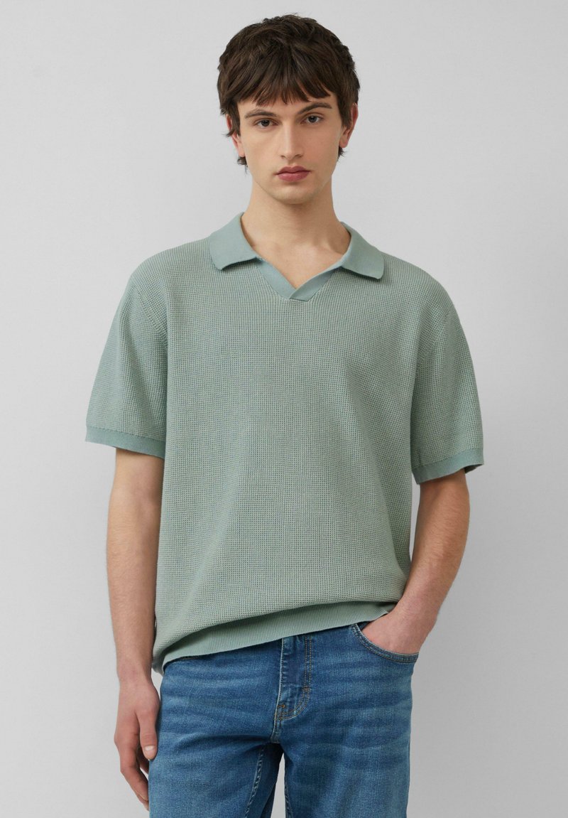 Polo à manches courtes, de couleur vert clair, en tissu texturé avec un décolleté en V et un col contrasté. Porté avec un jean bleu.