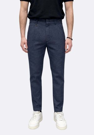 Uomo che indossa pantaloni blu scuro aderenti, camicia nera, sneakers bianche e un braccialetto nero di perline, in piedi con le mani lungo i fianchi.