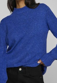 Blauwe gebreide trui met een geribde ronde hals, lange mouwen en een ontspannen pasvorm; gecombineerd met zwarte jeans. De textuur lijkt zacht en comfortabel.