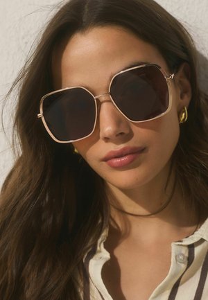 Woman with long brown hair wearing oversized square sunglasses, gold hoop earrings, and a striped buttoned shirt.