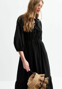 Black, long-sleeve dress with a tied neckline. Features a cinched waist and flowing skirt. Model holds a textured, round woven bag.