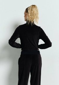 Kobieta z kręconymi blond włosami spiętymi w koński ogon, ubrana w czarną bluzkę z długim rękawem i spodnie, stoi z rękami na biodrach przy gładkiej białej ścianie.