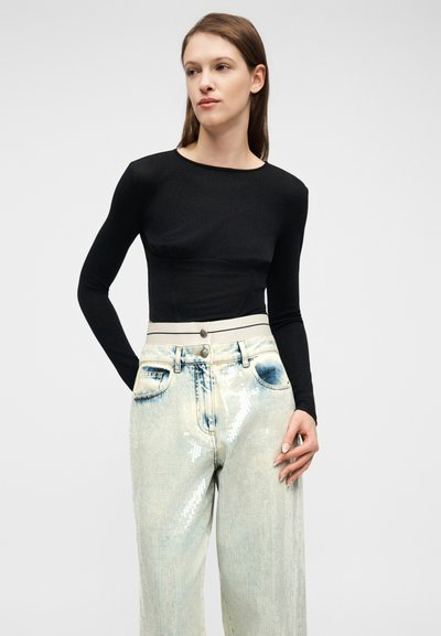 Γυναίκα με μακριά καστανά μαλλιά, ντυμένη με μαύρο εφαρμοστό μακρυμάνικο τοπάκι και τζην ψηλόμεσο ανοιχτού χρώματος με λαμπερό, αντανακλαστικό φινίρισμα.