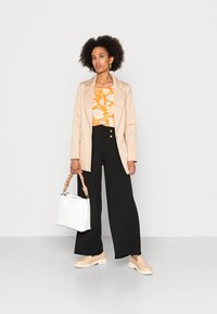Blazer bege sobre uma blusa floral laranja, calças largas pretas, mocassins de creme, segurando uma bolsa branca com alça trançada.