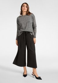 Grauer Strickpullover mit langen Ärmeln, der glänzende Akzente an den Manschetten aufweist. Schwarze, weit geschnittene Hose mit Bindegürtel. Schwarze, hochhackige Schuhe.
