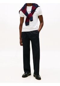 Camiseta blanca de manga corta con logo, pantalones plisados azul marino y un suéter a rayas azul marino y burdeos atado alrededor de los hombros. Mocasines negros.