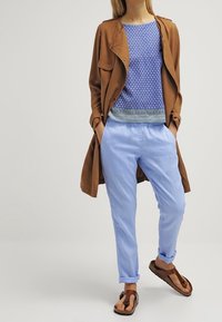 Abrigo largo marrón sobre blusa azul estampada, pantalones azul claro y sandalias marrones. Incluye un patrón geométrico y puños enrollados.