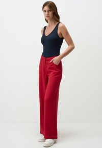 Czerwone spodnie typu wide-leg z wysokim stanem i kieszeniami z przodu, w zestawieniu z dopasowanym granatowym topem na ramiączkach. Gładka faktura, nowoczesny krój i białe trampki.