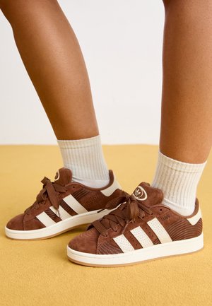Brązowe buty sportowe Adidas Campus z białymi paskami, noszone z białymi prążkowanymi skarpetami za kostkę na żółtej podłodze.