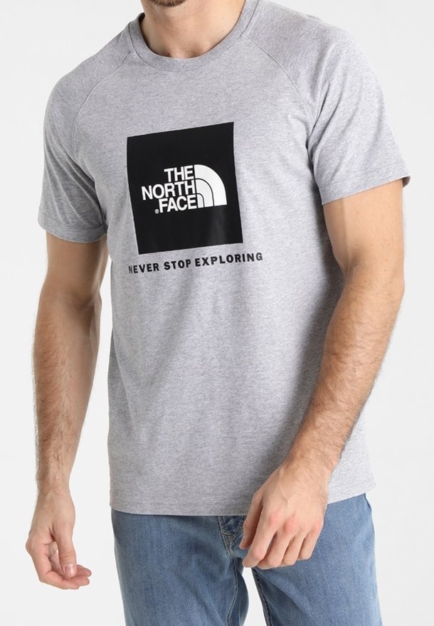 Camiseta de algodón gris con mangas cortas, que presenta un logo rectangular negro de "THE NORTH FACE" y el texto "NUNCA DEJES DE EXPLORAR" debajo.
