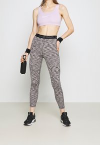 Sportovní podprsenka ve světle fialové, vysoké legíny se vzorem v šedých odstínech, černé logo Nike a černé sportovní boty. Lahev s vodou v ruce.
