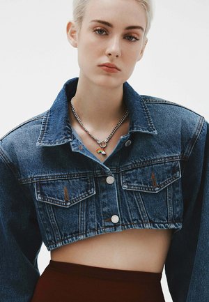 Chaqueta de denim azul cortada con dos bolsillos frontales, cierres de botón plateados, combinada con un collar de cadena plateada que presenta un pequeño encantamiento de arcoíris.
