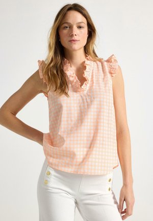 Kvinde med lysebrunt hår iført en ferskenfarvet ternet ærmeløs bluse med rynket halsudskæring og hvide bukser med guldknapper.