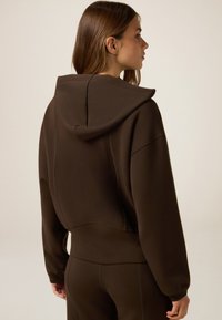 Felpa marrone con un design cropped, caratterizzata da un ampio cappuccio e dettagli di cucitura visibili. Il tessuto appare liscio e morbido.