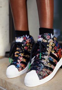 Zwarte hoge sneakers met een meerkleurig textuurpatroon, voorzien van een witte rubberen neuskap en zwarte veters, met groene veteruiteinden als accent.