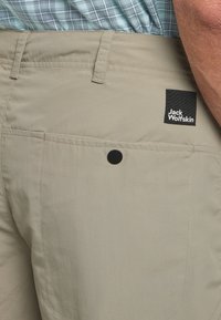 Pantaloni corti beige con una trama liscia. Presentano un'etichetta nera con il logo "Jack Wolfskin" e una tasca posteriore con bottone.