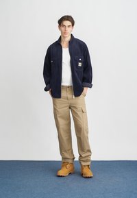 Camisa de corduroy azul-marinho com dois bolsos no peito, t-shirt branca, calças cargo bege e botins de trabalho castanhos com atacadores sobre um tapete azul.