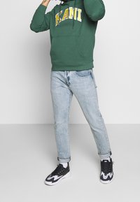 Grön hoodie med "KANI" i gult och vitt, ljusblå jeans och svart-vita sneakers. Hoodien har en framficka och dragsnören.