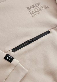 Beige kapuutsiga särk tekstuuri viimistlusega, musta tõmbluku sulgemise ja etiketi sildiga. "BAKER by TED BAKER" on paisutatud tumedamas varjus.