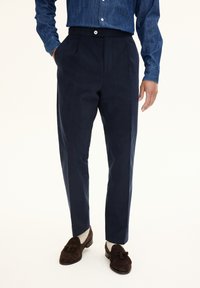 Navy trousers with a tailored fit, featuring two front pleats, a button closure, and side pockets, paired with brown suede loafers.