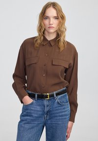 Brązowa koszula z długim rękawem i dwoma kieszeniami na piersi, gładka w dotyku, w połączeniu z niebieskimi dżinsami o wysokim stanie i czarnym, teksturowanym paskiem.