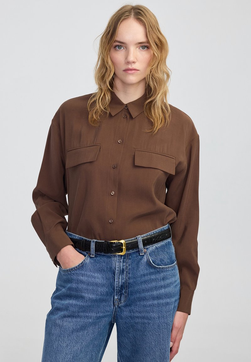 Brązowa koszula z długim rękawem i dwoma kieszeniami na piersi, gładka w dotyku, w połączeniu z niebieskimi dżinsami o wysokim stanie i czarnym, teksturowanym paskiem.