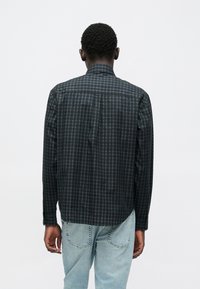 Mustast ja hallist ruuduline särk nööbitava krae, pikkade varrukate ja sirge äärega. Tagantvaates on näha peeneid detaile. Kerge sinine teksapüksid on nähtavad.