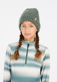 Groen gebreid beanie met een gestructureerd patroon en een klein leren label. Ggedragen met een langemouwen top met rits in aquakleurige strepen.
