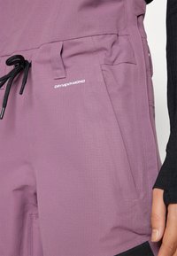 Lilla tekstuuriga kangas, millel on nööriga vöökoht, küljetasku ja valge "DRYVENT-MONO" logo. Mustad aktsendid all.