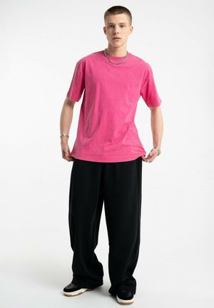 Młody mężczyzna ubrany w jasnoróżową, oversize'ową koszulkę, luźne czarne spodnie, czarne sneakersy oraz srebrny naszyjnik, stoi na tle białego tła.
