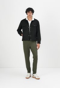 Chaqueta negra con un cuello blanco, dos bolsillos en el pecho y botones plateados. Pantalones verde oliva y zapatillas blancas con suelas marrones.