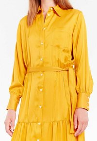 Robe jaune ceinturée en matériau lisse. Présente des manches longues, un devant à boutons, une poche poitrine et une jupe à volants avec des poignets boutonnés.