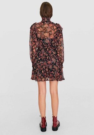 Woman standing facing away wearing a sheer black floral dress with long sleeves and red ankle boots on a plain light background.