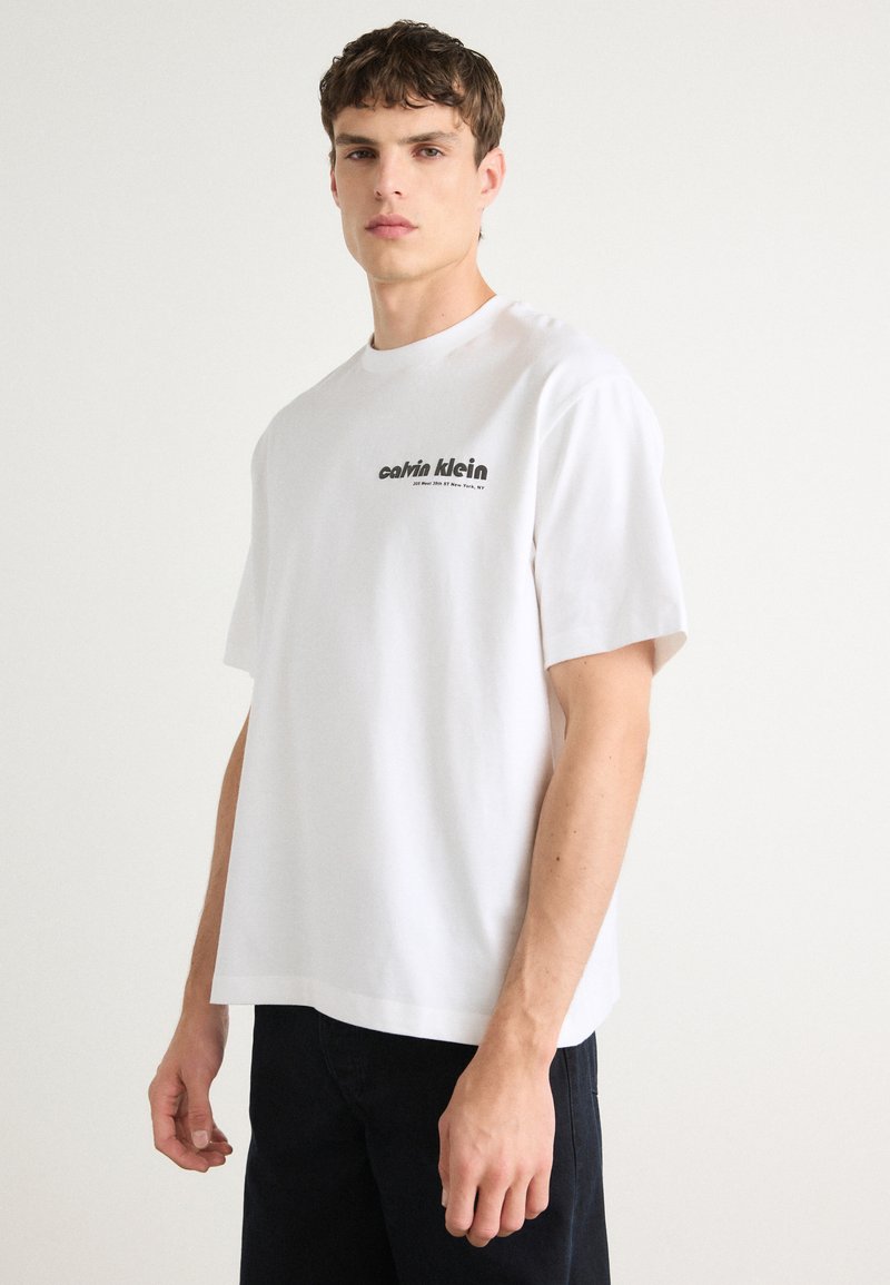 Balta kokvilnas T-kreklu ar brīvu piegriezumu, īsām piedurknēm, apaļu izgriezumu un ar uzdrukātu "Calvin Klein" logotipu kreisajā pusē.